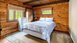 Une chambre à coucher dans une cabane en bois, avec un lit, deux fenêtres équipées de stores, une armoire et une lampe de chevet.