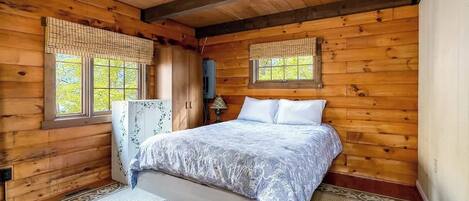 Une chambre à coucher dans une cabane en bois, avec un lit, deux fenêtres équipées de stores, une armoire et une lampe de chevet.