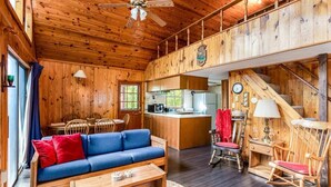 Interior de una cabaña de madera con sofá, mesa de comedor, sillas, zona de cocina y una escalera.