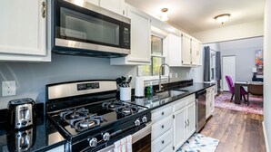 Una cucina moderna con piano cottura a gas, forno a microonde ed elettrodomestici in acciaio inossidabile. Si vede una finestra con tende, un lavello e un'area pranzo con una sedia.