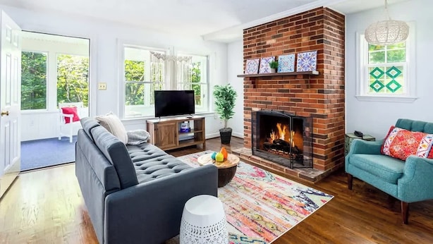 A living room with a brick fireplace, a sofa, a television, and a window with a view of greenery. - Mountain home w/ hot tub & sunroom (Front Royal)