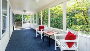 A covered porch with white chairs and a table, large windows offering a view of greenery. - Mountain home w/ hot tub & sunroom (Front Royal)