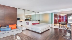 A modern hotel room with a large bed, a sofa, a glass coffee table, and a dining area with a chair.