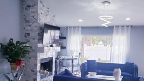 A modern living room with a blue sofa, a flat-screen TV, a fireplace, and a dining area with glass furniture. - Bright & Airy St. Pete Home w/ Outdoor Living (St. Petersburg)