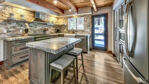 A modern kitchen with a large island, wooden ceiling beams, and a stone backsplash.