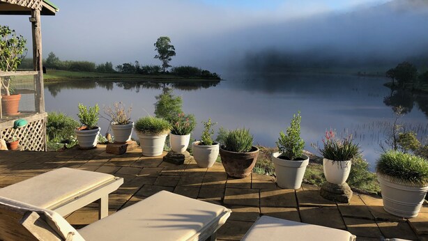A serene lakeside patio with lounge chairs and potted plants, overlooking a calm lake and misty hills. - Historic Georgian Stone Built Cottage (Saint Albans)