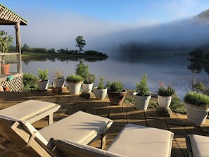 Un patio tranquillo affacciato su un lago calmo, con poltrone da relax e piante in vaso, e vista su dolci colline nebbiose.