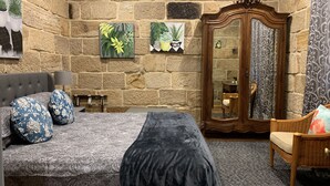 A bedroom with a stone wall, a bed with patterned bedding, a mirror, and a chair. - Historic Georgian Stone Built Cottage (Saint Albans)