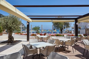 Un patio al aire libre con mesas y sillas de mimbre, una pérgola y vistas al mar y sombrillas de playa.