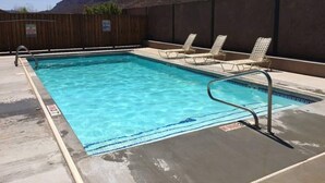 Piscina retangular com água azul cristalina, cercada por uma vedação de madeira e um deck de concreto. Existem cadeiras de descanso ao longo da borda da piscina.
