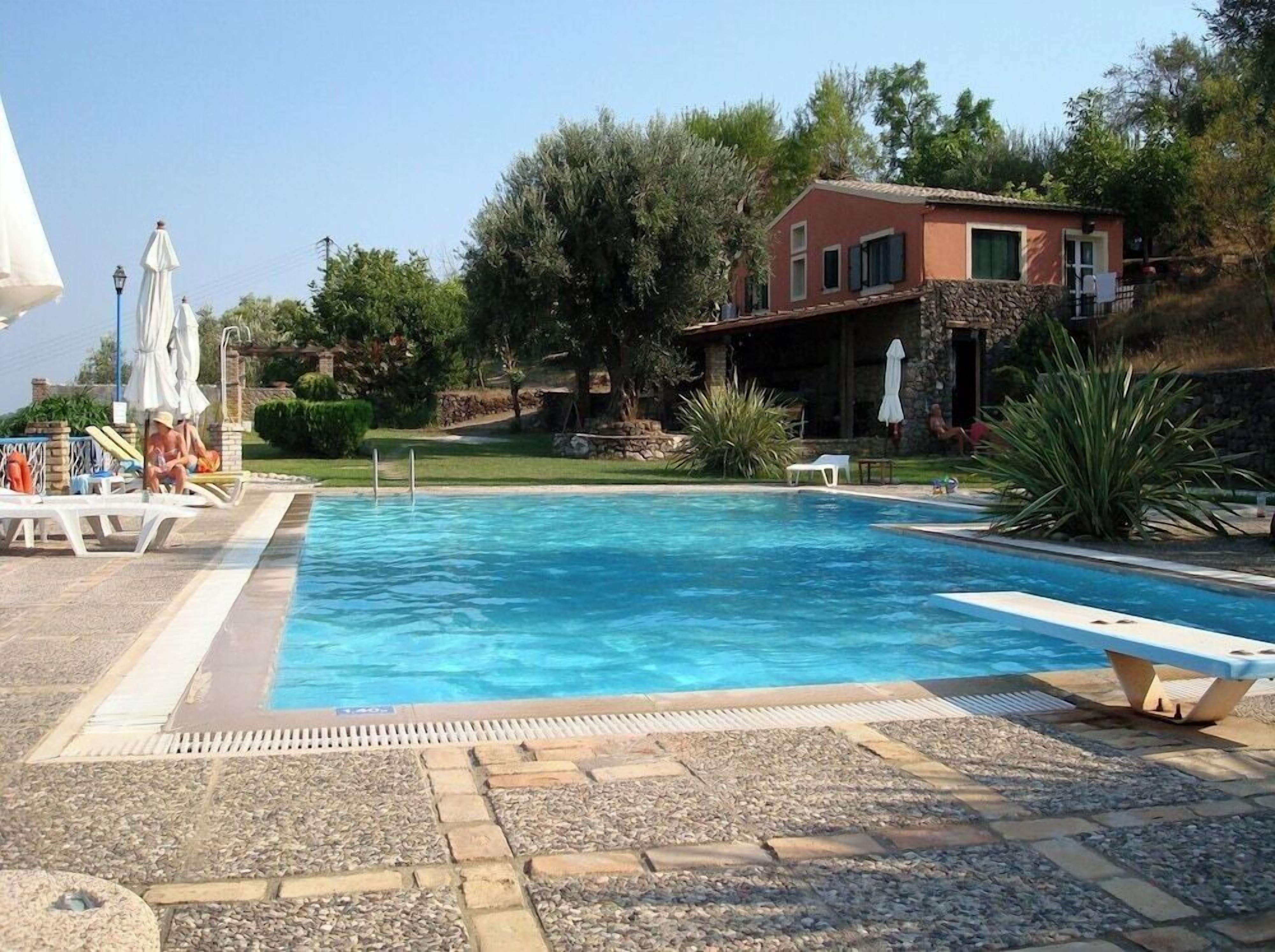 A swimming pool with lounge chairs and umbrellas, surrounded by greenery and a stone building.