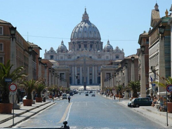 A street leading to a grand building with a large dome, flanked by historic buildings and palm trees. - Saint Peter Six Rooms And Suites (Rome)