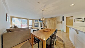 A modern living room with a sofa, a dining table with chairs, a chandelier, and a kitchen area with a refrigerator and cabinets. - Liftside 205 (Keystone)