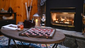 A cozy room with a wooden wall, a fireplace, a chessboard, and a candle on a table. - Schroon River Retreat, Waterfront, Near Lake George & Gore Mountain, Hot tub (Warrensburg)