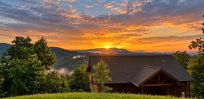 Un chalet rustique avec un toit en pente, entouré d’une végétation luxuriante et dominé par un paysage montagneux au coucher du soleil.