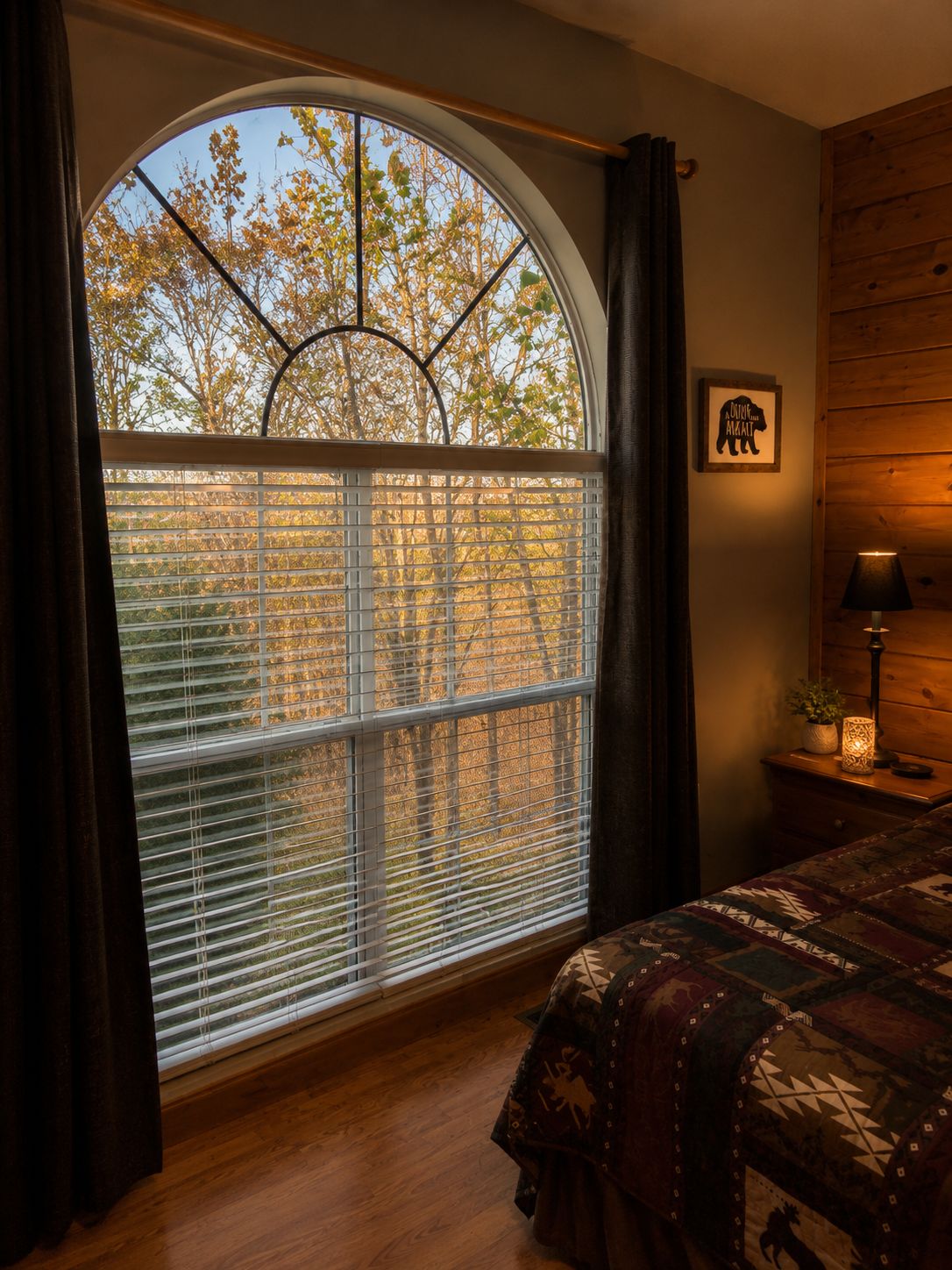 A bedroom with a large arched window, a bed with patterned bedding, a bedside table with a lamp, and a framed picture on the wall.