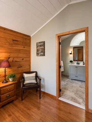 Una habitación con piso y pared de madera, una silla con cojín, una mesita de noche con lámpara, una planta en maceta y un cartel en la pared.