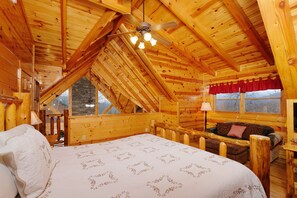 Een slaapkamer in een blokhut met een bed, een houten plafondventilator, een raam met een rode gordijn en een stenen muur die door het raam zichtbaar is.