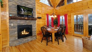 L’intérieur chaleureux d’un chalet avec une cheminée en pierre, une table à manger en bois et un téléviseur fixé au mur.