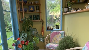 Una habitación acogedora con un amplio ventanal, sillas de mimbre, plantas en macetas y estantes con adornos.