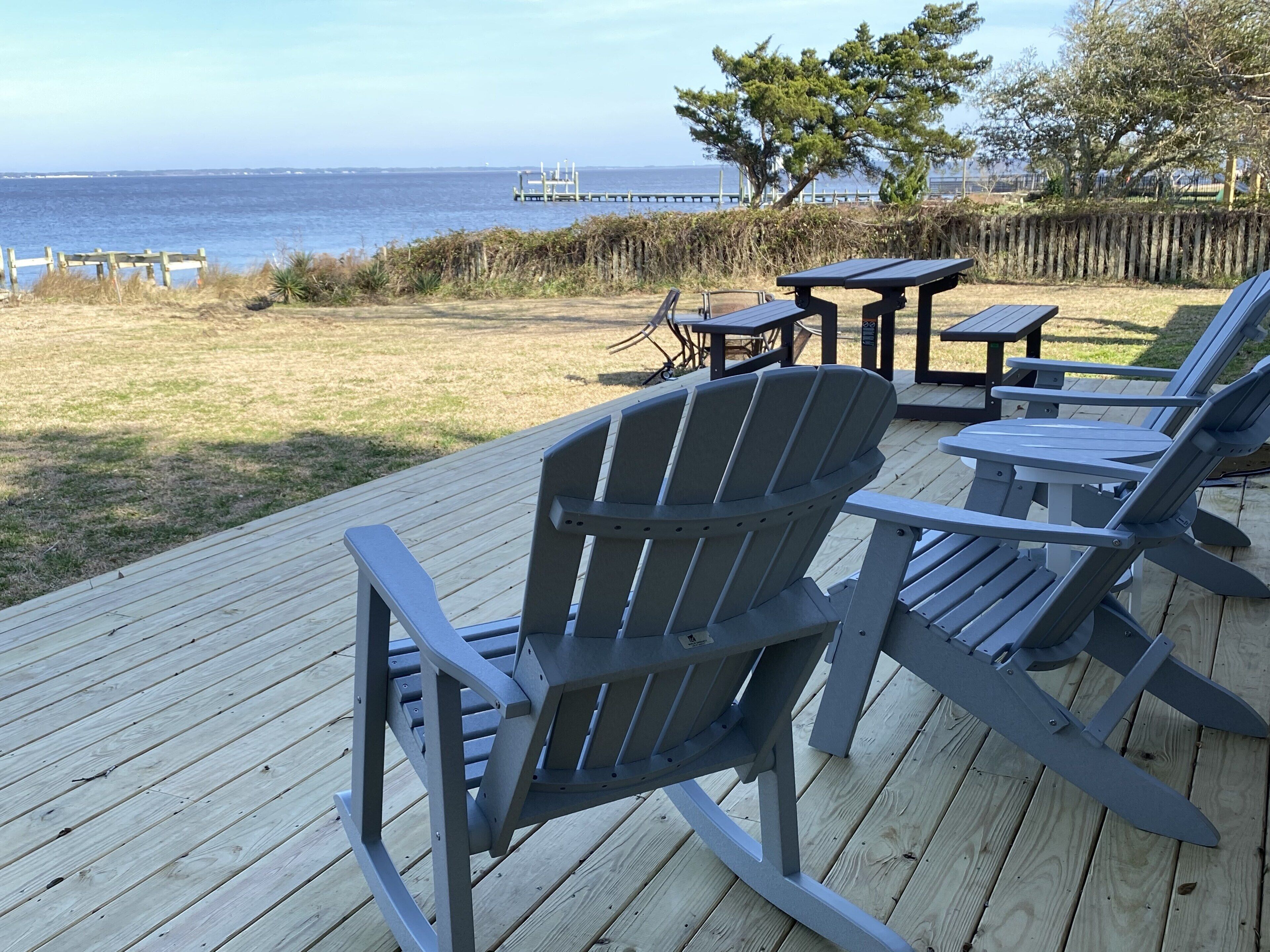 A deck with Adirondack chairs and a table, overlooking a grassy area and the ocean.