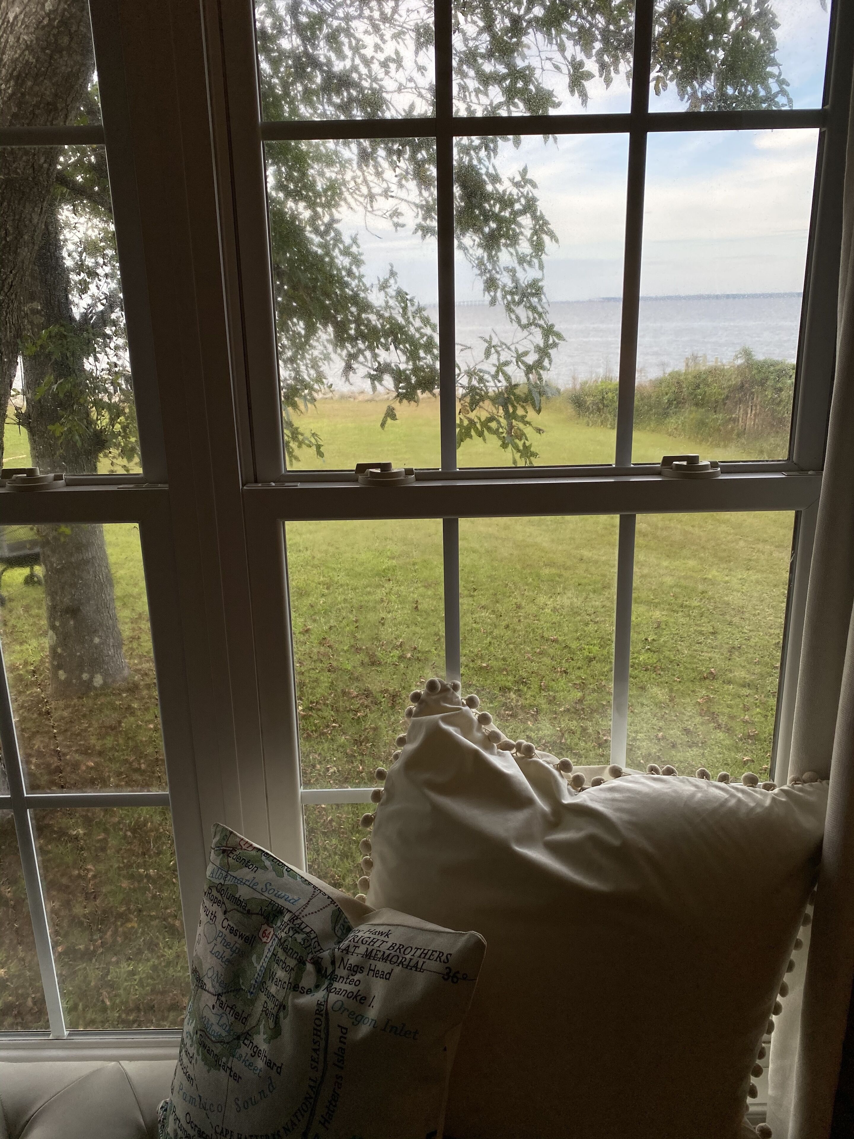 Une fenêtre donnant sur un espace verdoyant et des arbres, un coussin orné d’un motif textuel et un oreiller blanc posé sur une chaise.