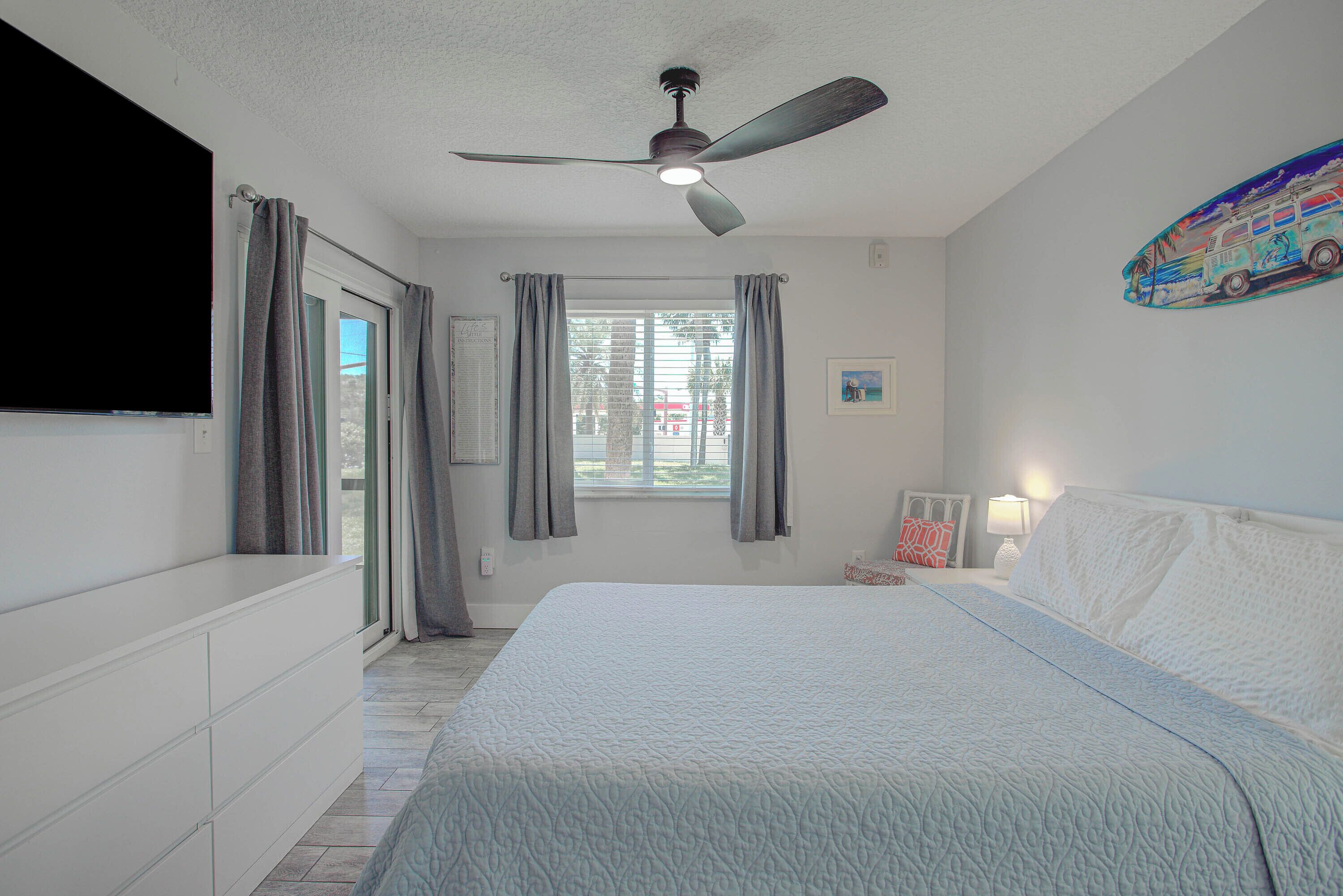 Un dormitorio con cama, ventilador de techo, ventana con cortinas, televisor montado en la pared y un marco de fotos.