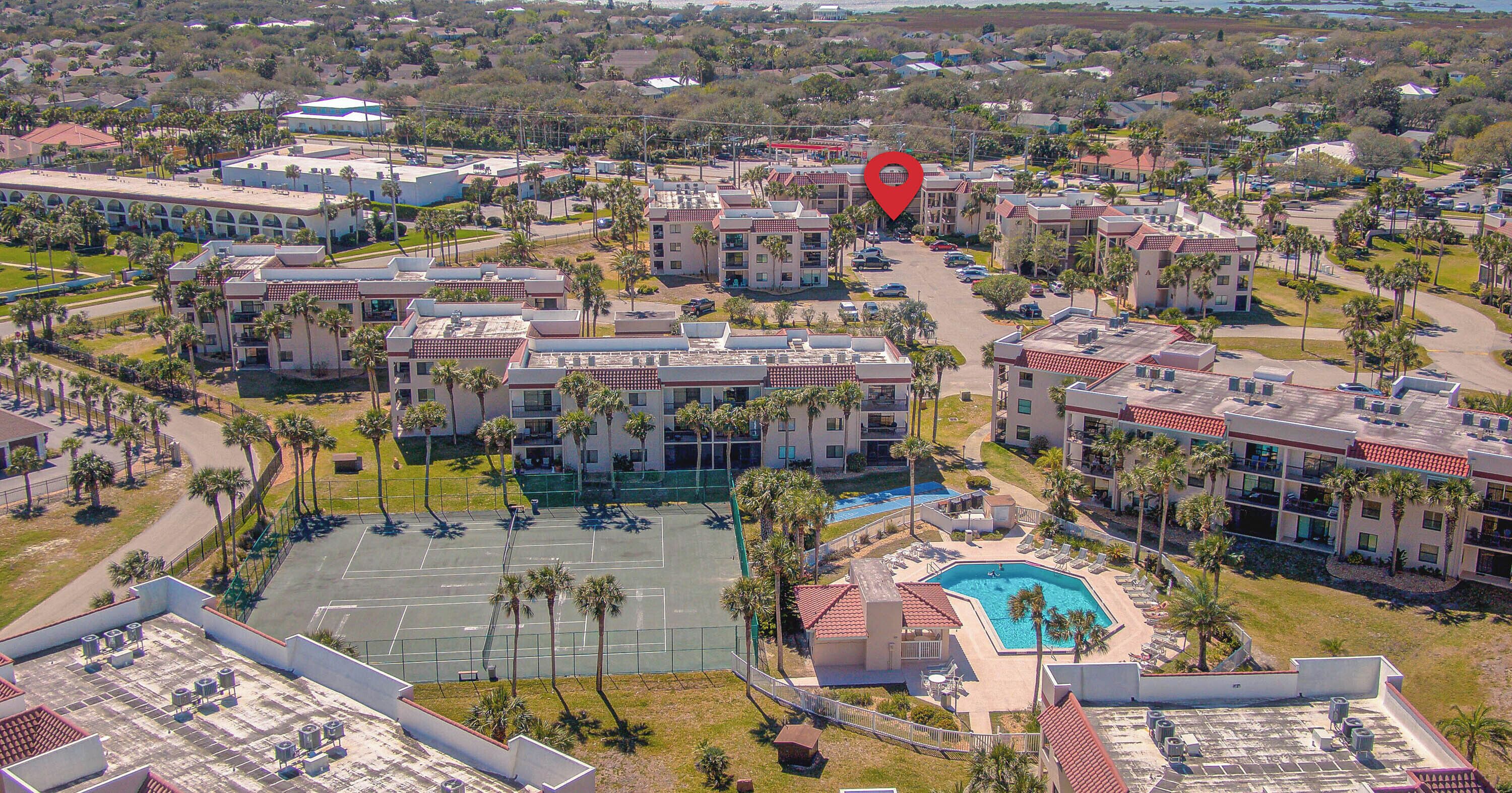 Vista aérea de un complejo turístico con varias edificaciones, una piscina y una cancha de tenis.
