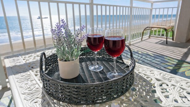 A wicker table with two wine glasses and a potted plant on a balcony overlooking the ocean.