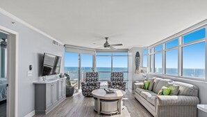 A spacious living area with a large window overlooking the ocean, a ceiling fan, and a seating arrangement including a sofa and chairs.