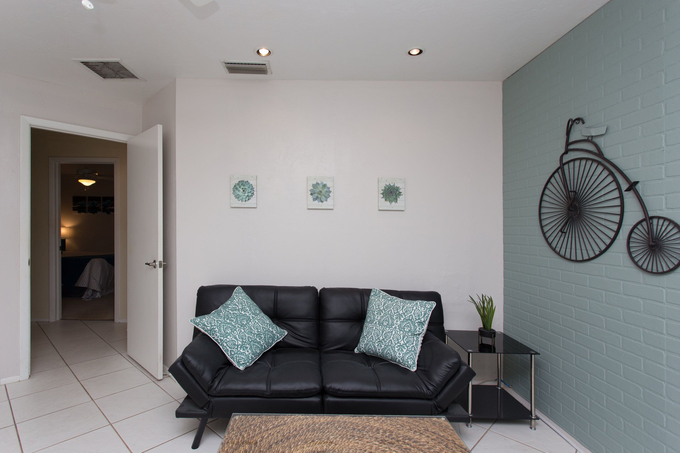 A modern living room with a black leather sofa, teal patterned cushions, and a coffee table. There are three wall-mounted decorative items and a large bicycle decoration on the wall.
