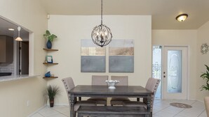 Een eetruimte met een donker houten tafel, beige stoelen en een kroonluchter. Boven de tafel hangen twee wanddecoraties en links staat een potplant.