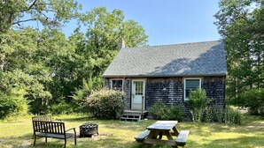 A small wooden cabin with a porch, a picnic table, and a bench in a grassy yard surrounded by trees. - Private cottage nestled in center of the Vineyard (West Tisbury)