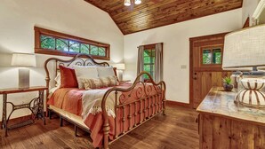 Un dormitorio con techo de madera, una cama con estructura metálica, un aparador de madera y un ventanal con cortinas.