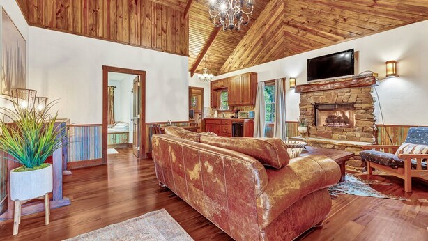 Un salón amplio con un sofá de cuero grande, piso de madera, una chimenea de piedra y un televisor montado en la pared.