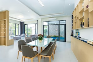 Um interior moderno com uma mesa de jantar de vidro, cadeiras estofadas cinzas, uma estante de madeira e vista para um jardim através de grandes janelas.