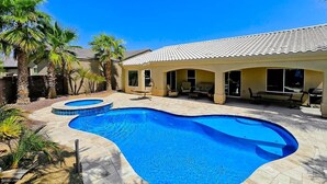 A swimming pool with a curved design, surrounded by palm trees and a patio area with outdoor furniture. - Cozy Island House with Heated Pool (Lake Havasu City)