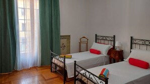 Een kamer met twee eenpersoonsbedden, een houten vloer, een raam met groene gordijnen en een kroonluchter.