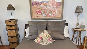 A bed with a grey bedspread and multiple pillows, a wicker storage unit, a framed painting, and a small table with a lamp and candle. - Casa de San Pasquale, 1800s restored terones adobe (Albuquerque)