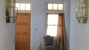 A room with wooden flooring, a wooden door, two windows with curtains, a chair, and two wall-mounted lanterns. - Casa de San Pasquale, 1800s restored terones adobe (Albuquerque)
