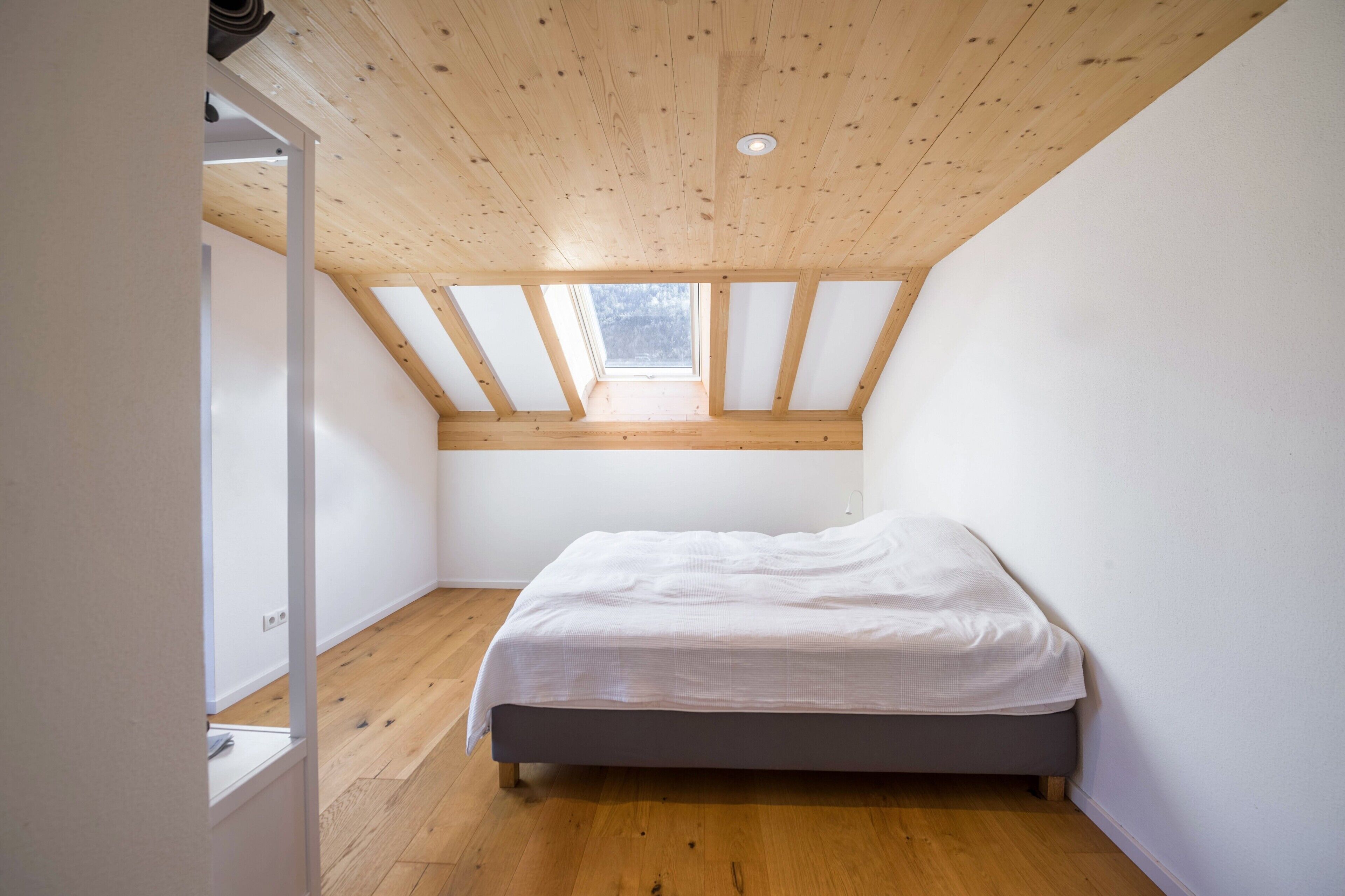 Un dormitorio con techo inclinado, vigas de madera, una cama individual y un tragaluz.