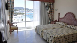 Habitación de hotel con una cama grande, una silla, una mesita de noche y vistas a una playa y edificios a través del ventanal.