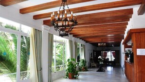 Un pasillo amplio con vigas de madera, ventanas grandes con cortinas y plantas en macetas.