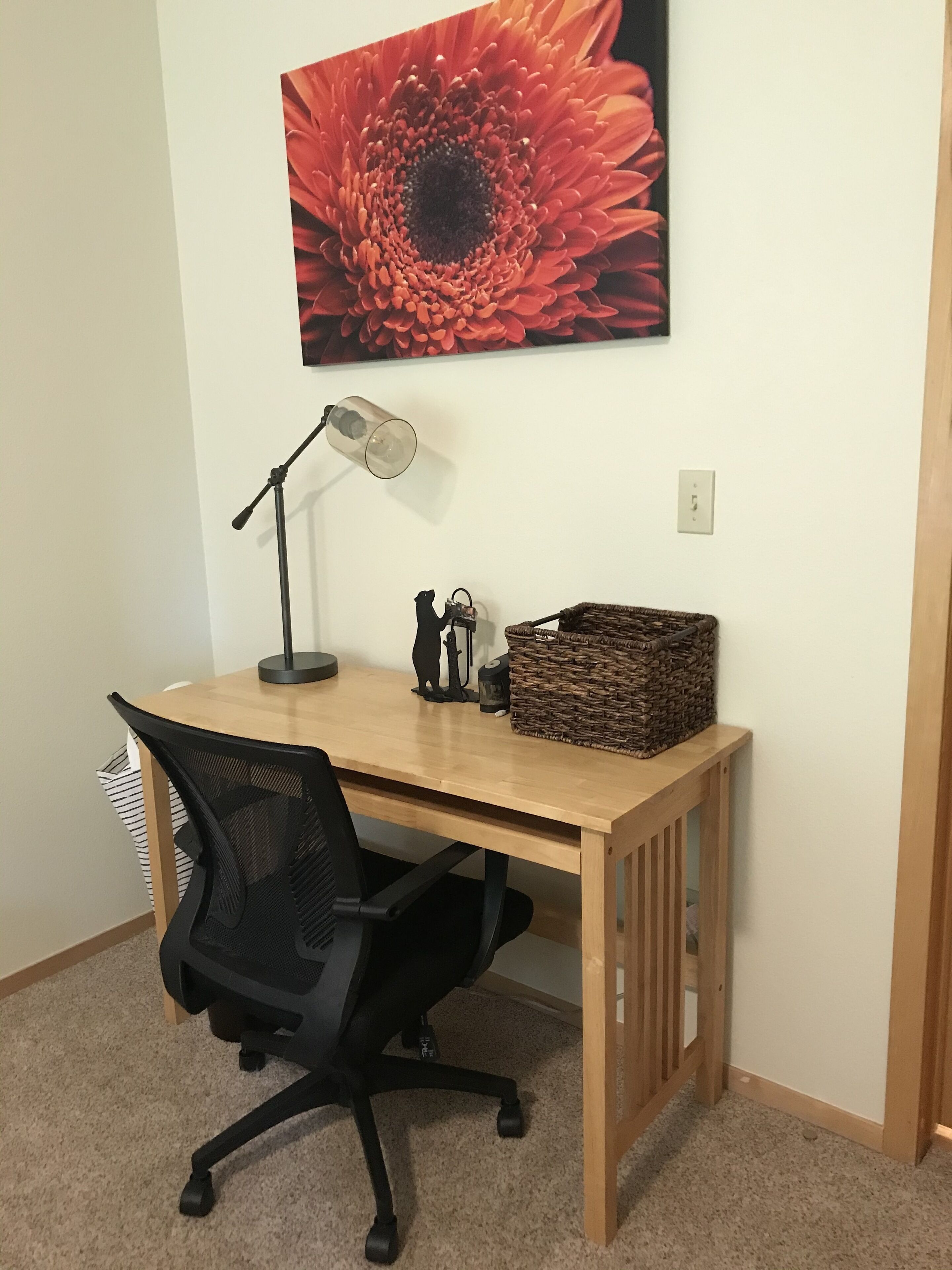 A wooden desk with a black office chair, a lamp, a basket, and a bear figurine.