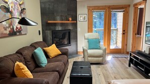 A modern living room with a brown leather sofa, teal and mustard cushions, a black ottoman, a wooden chair with a teal cushion, a fireplace, and a television.