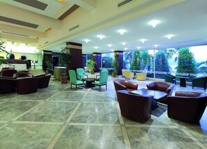 Un'ampia hall con reception, diverse zone salotto con poltrone in pelle e tessuto, e ampie finestre che offrono vista sul verde circostante.
