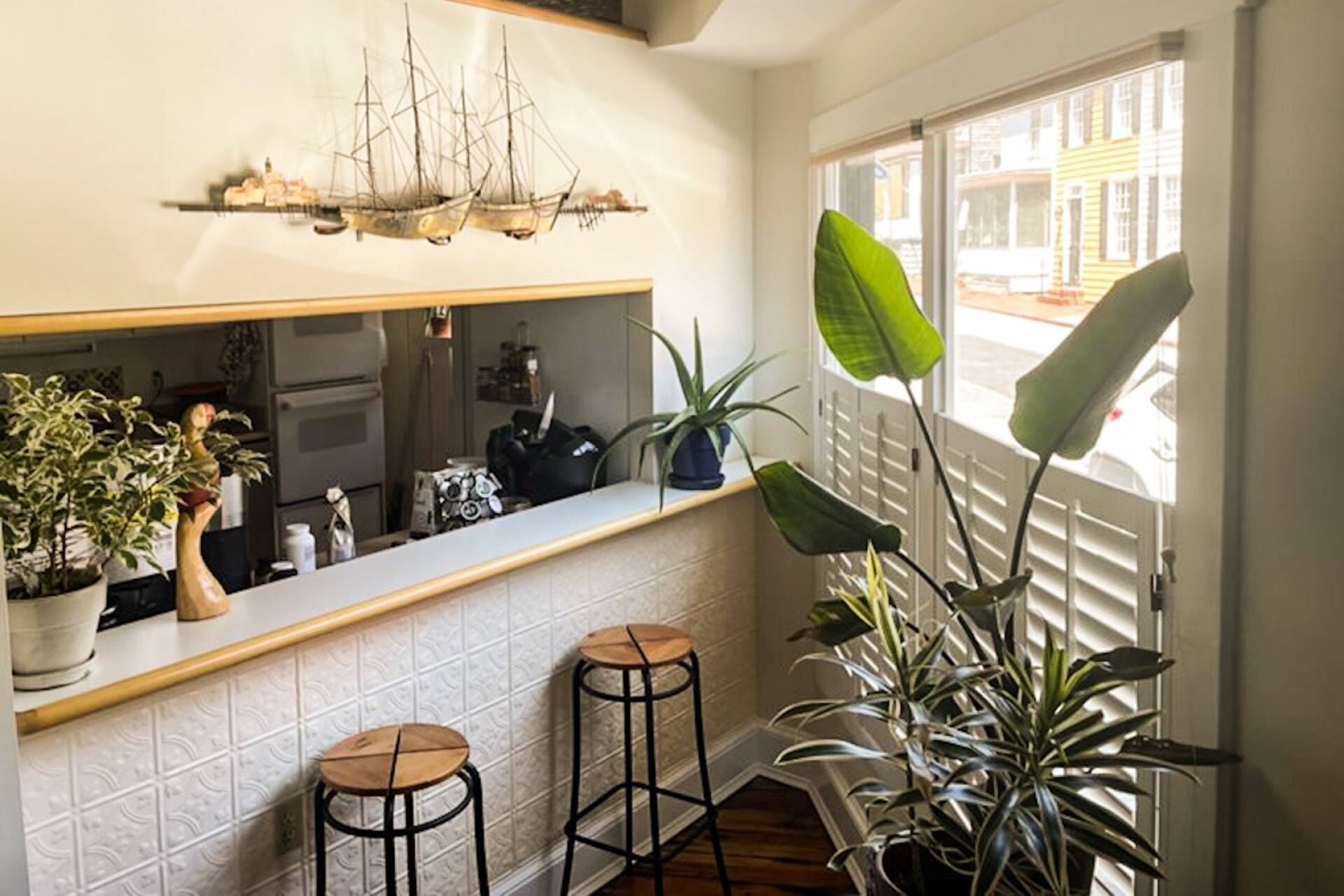 A kitchen with a tiled backsplash, a ship-themed artwork, two stools, and potted plants.