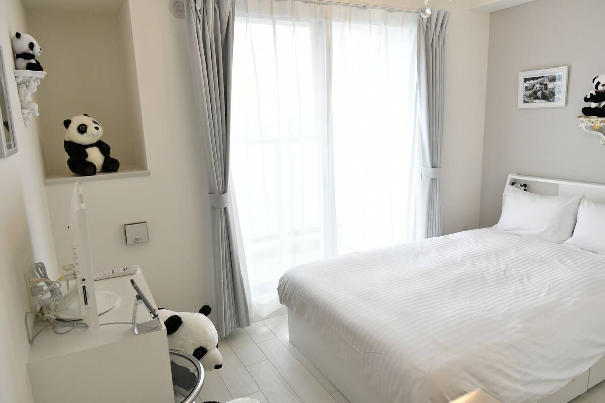 A bedroom with a bed, a desk with a computer, a panda plush toy, and a window with curtains.