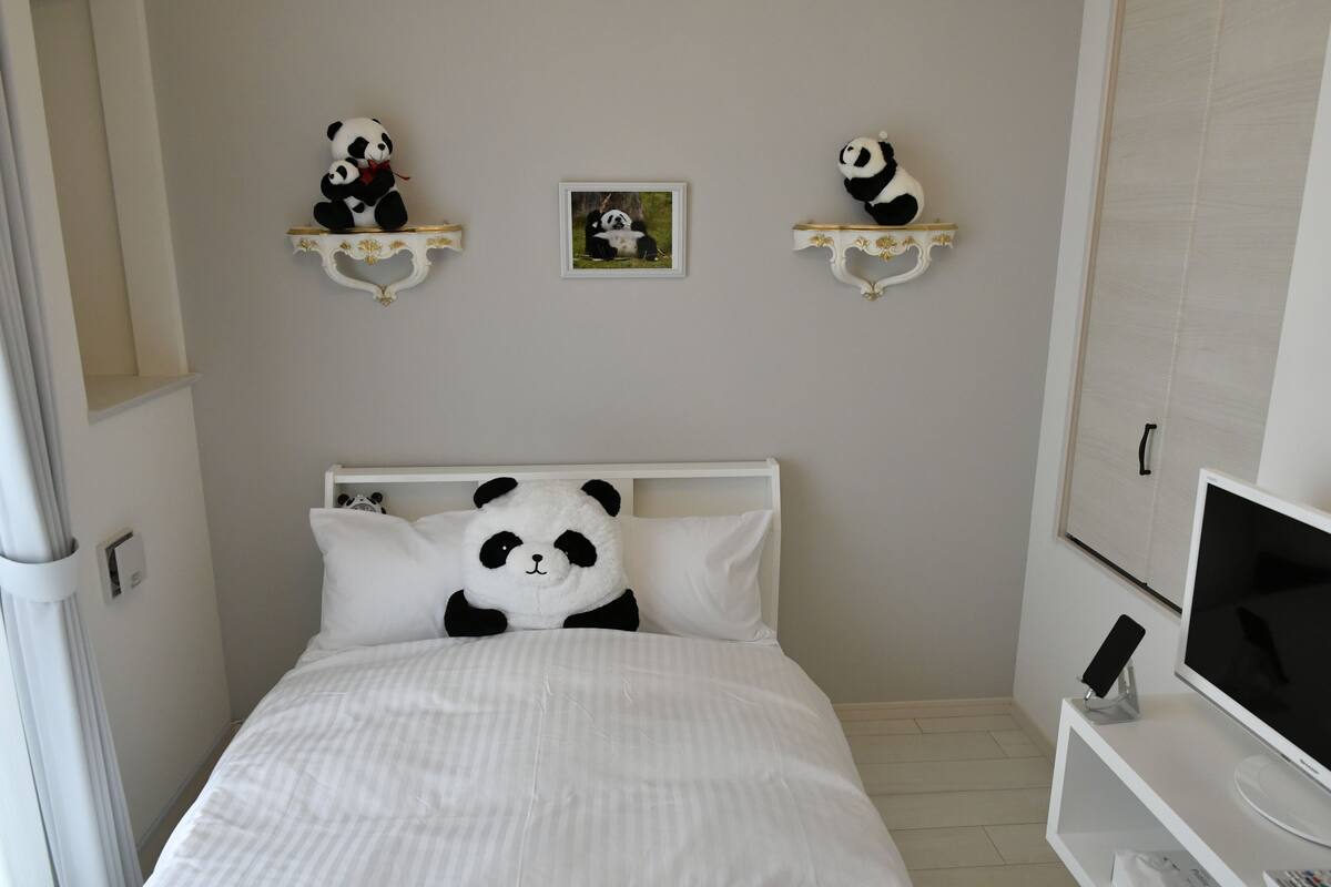 A neatly arranged bedroom with a bed, a TV on a stand, and two panda-themed wall decorations.