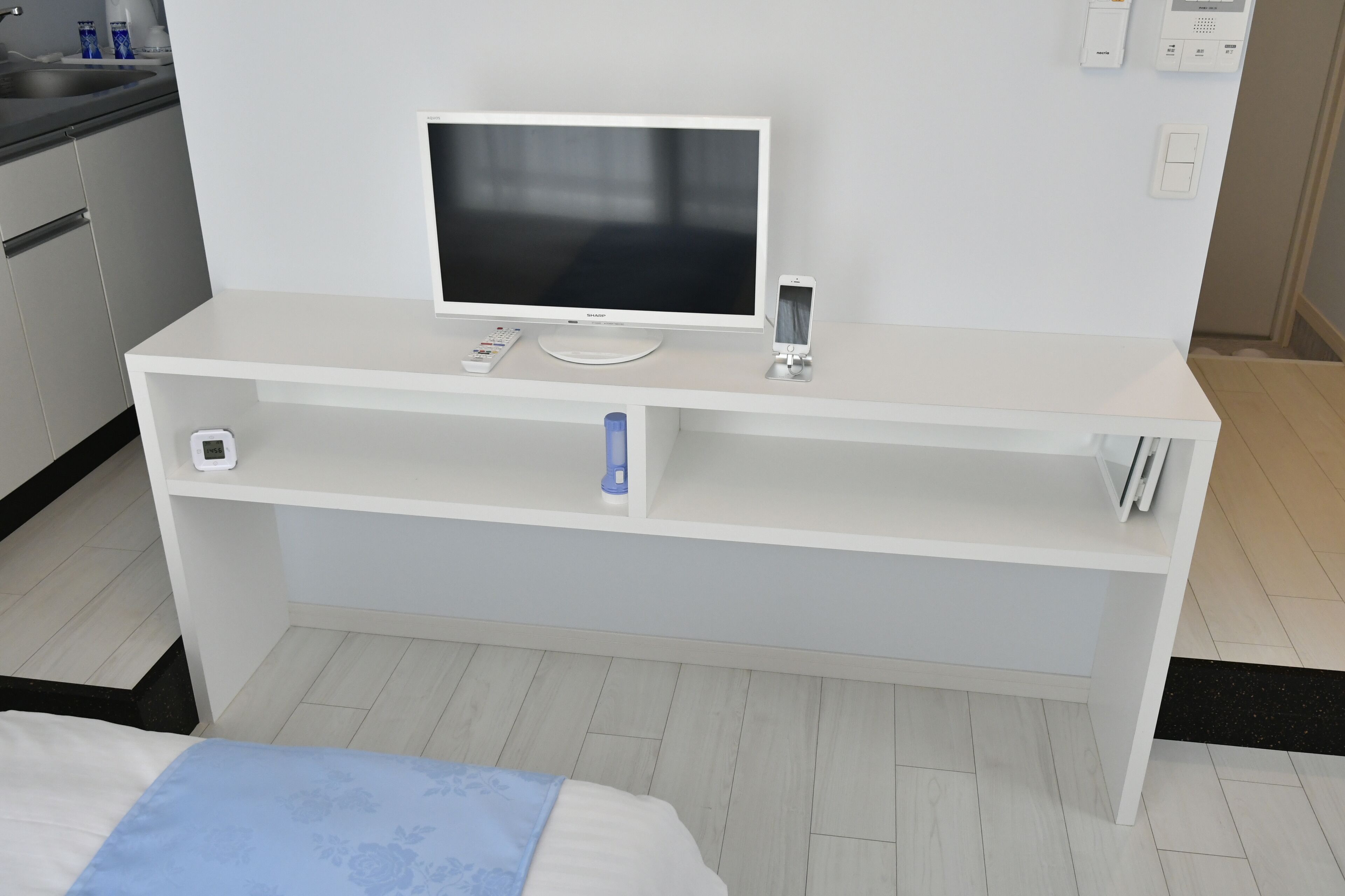 A modern hotel room with a white TV on a stand, a small bedside table, and a drawer unit below.